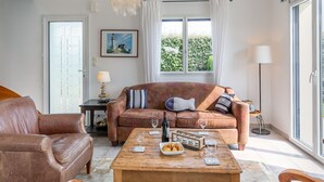 Una sala de estar con un sofá de cuero marrón, una mesa de centro de madera con copas de vino y aperitivos, una lámpara colgante y vistas a la vegetación exterior.