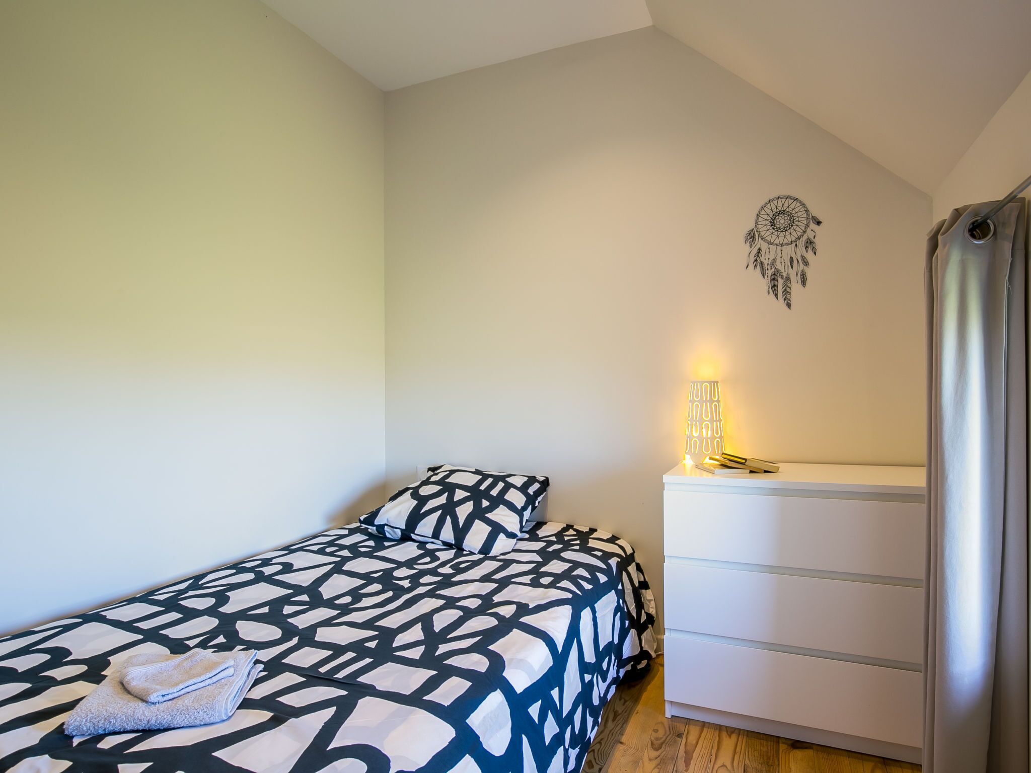 Un dormitorio pequeño y ordenado con una cama, una mesita de noche con una lámpara, un aparador y un atrapasueños en la pared.