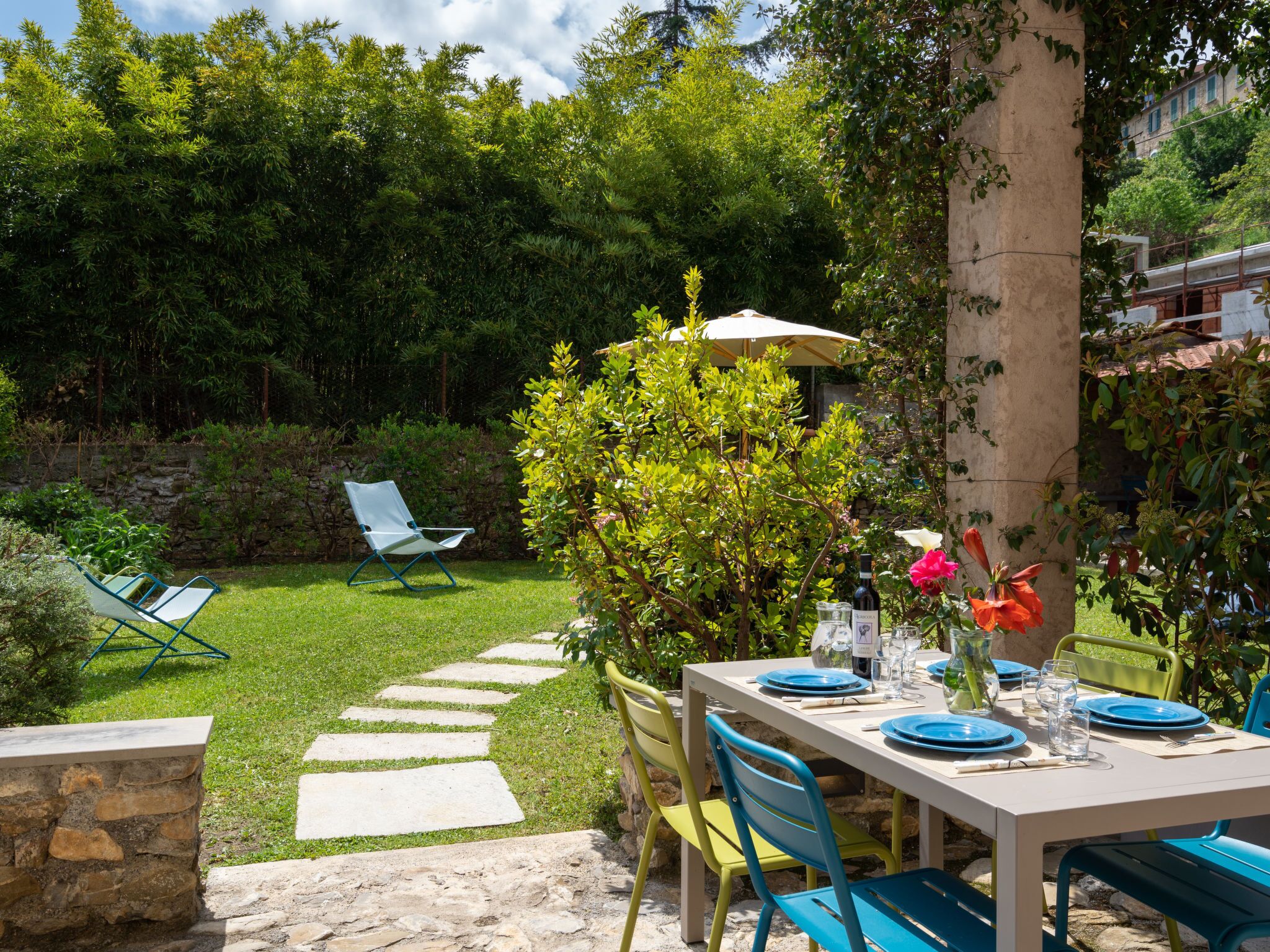 An outdoor dining area with a table set for two, surrounded by greenery and a stone pathway.