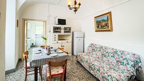 Een kleine woonruimte met een eettafel, stoelen, een bank, een kitchenette en een hanglamp.