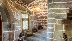 Un intérieur aux murs de pierre, avec une porte en bois, une fenêtre ornée d’une grille décorative et un petit escalier menant à un niveau supérieur.