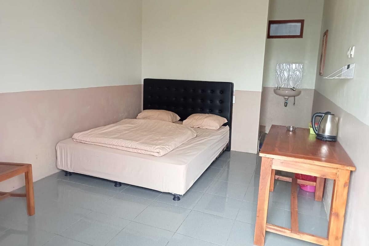 Uma cama de solteiro com cabeceira preta num quarto com piso de azulejos, uma mesa de madeira e uma pia pequena.