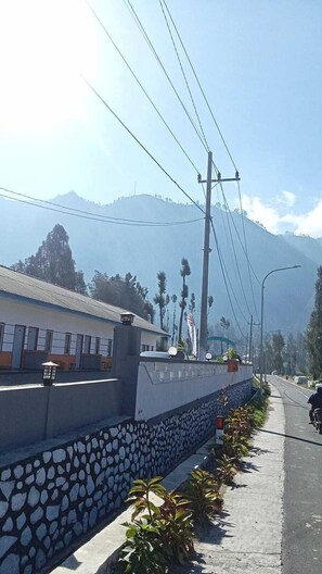 A roadside view with a stone wall, a building with a corrugated metal roof, and a mountain in the background. - Tengger Indah Seruni Bromo (Ngadisari)