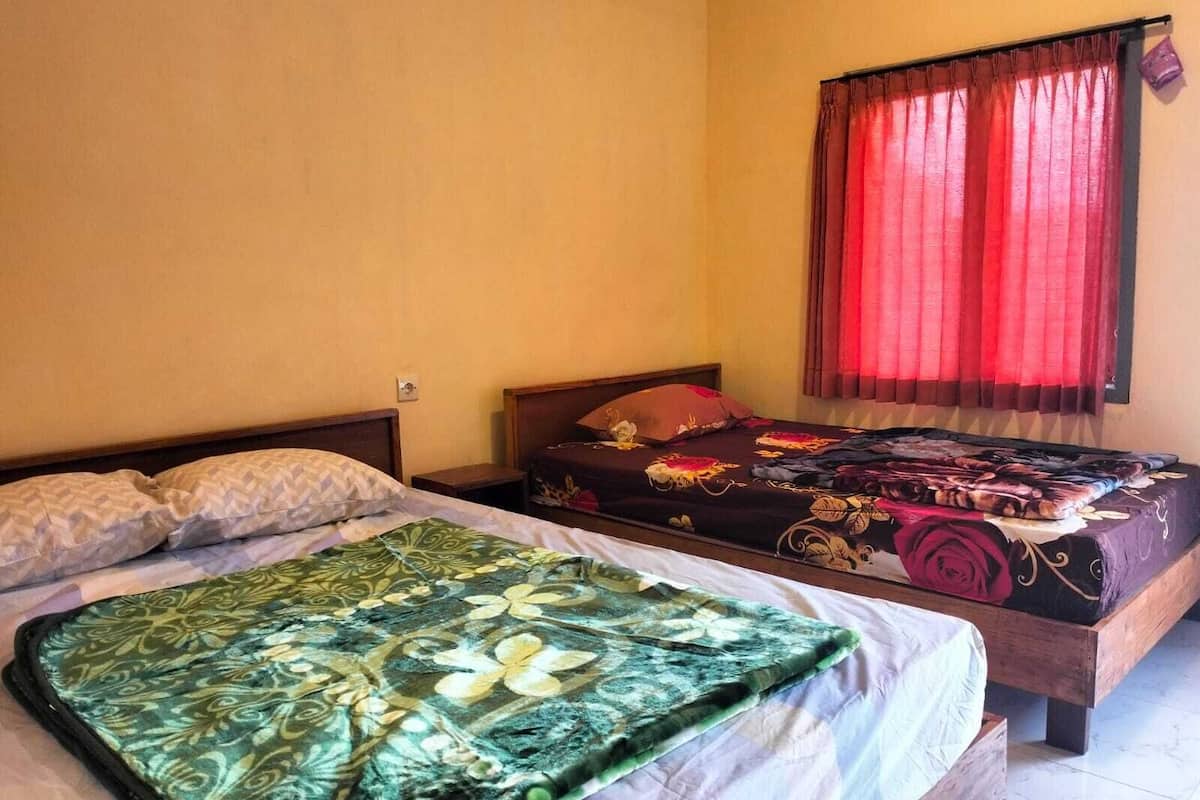Quarto com duas camas, uma com colcha florida verde e a outra com colcha florida escura. Há uma janela com cortinas vermelhas e uma tomada visível.