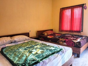 A room with two beds, one with a green floral bedspread and the other with a dark floral bedspread. There is a window with red curtains and a wall socket visible. - Tengger Indah Seruni Bromo (Ngadisari)