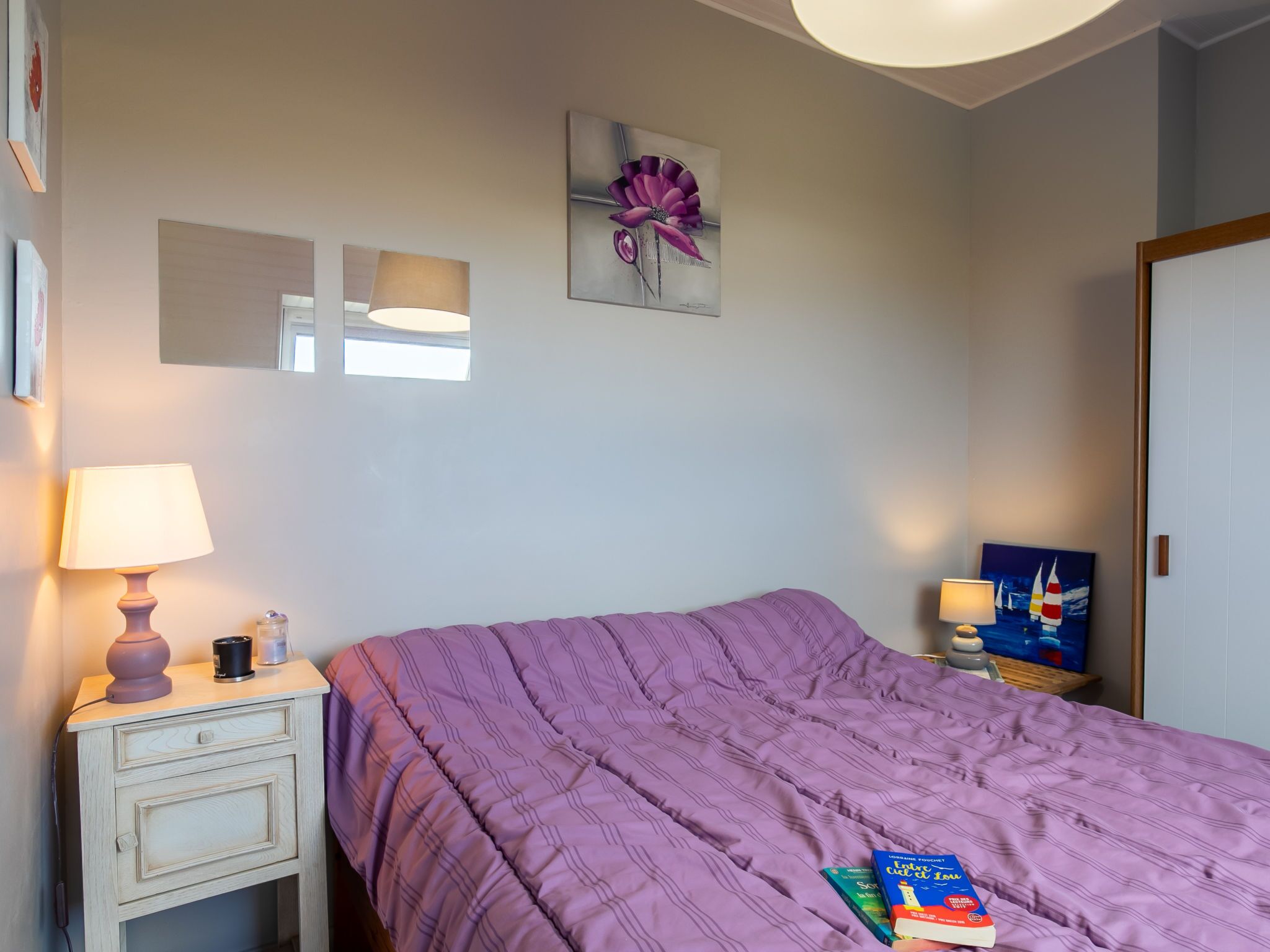 Ein Schlafzimmer mit violetten Bettwäsche, einer Nachttischlampe, einem kleinen Tisch mit Lampe und einem Blumenbild an der Wand.