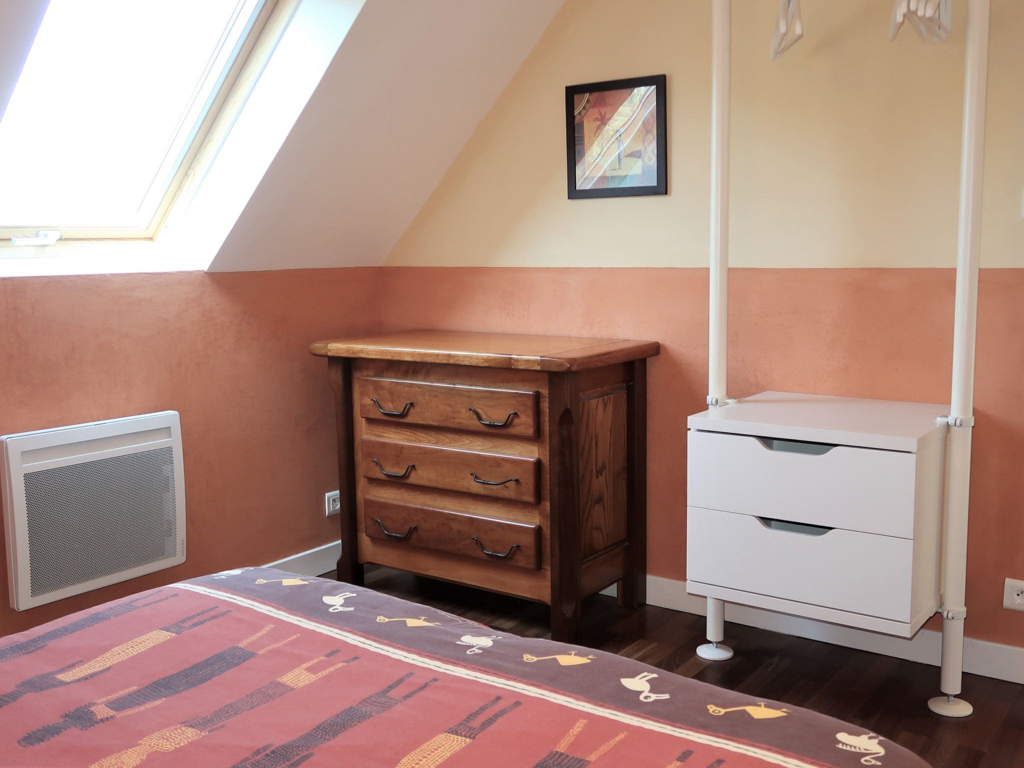 Une pièce avec un plafond en pente, une commode en bois avec des tiroirs, une armoire blanche, un radiateur et un tapis à motifs.