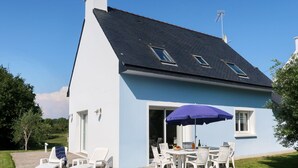 A two-story house with a blue exterior and a black roof. There is a patio with white chairs and a table, and a purple umbrella. - Ty Coat Pin by Interhome (Tregunc)