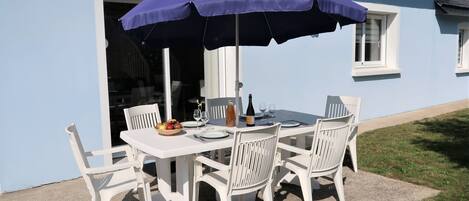 A table set outdoors with a purple umbrella, white chairs, and a bowl of fruit.