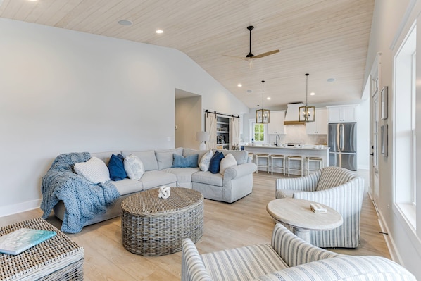 Un vaste salon comprenant un grand canapé, un pouf rond en osier et des fauteuils à rayures. On aperçoit à l’arrière-plan une cuisine équipée d’appareils modernes et un coin repas.