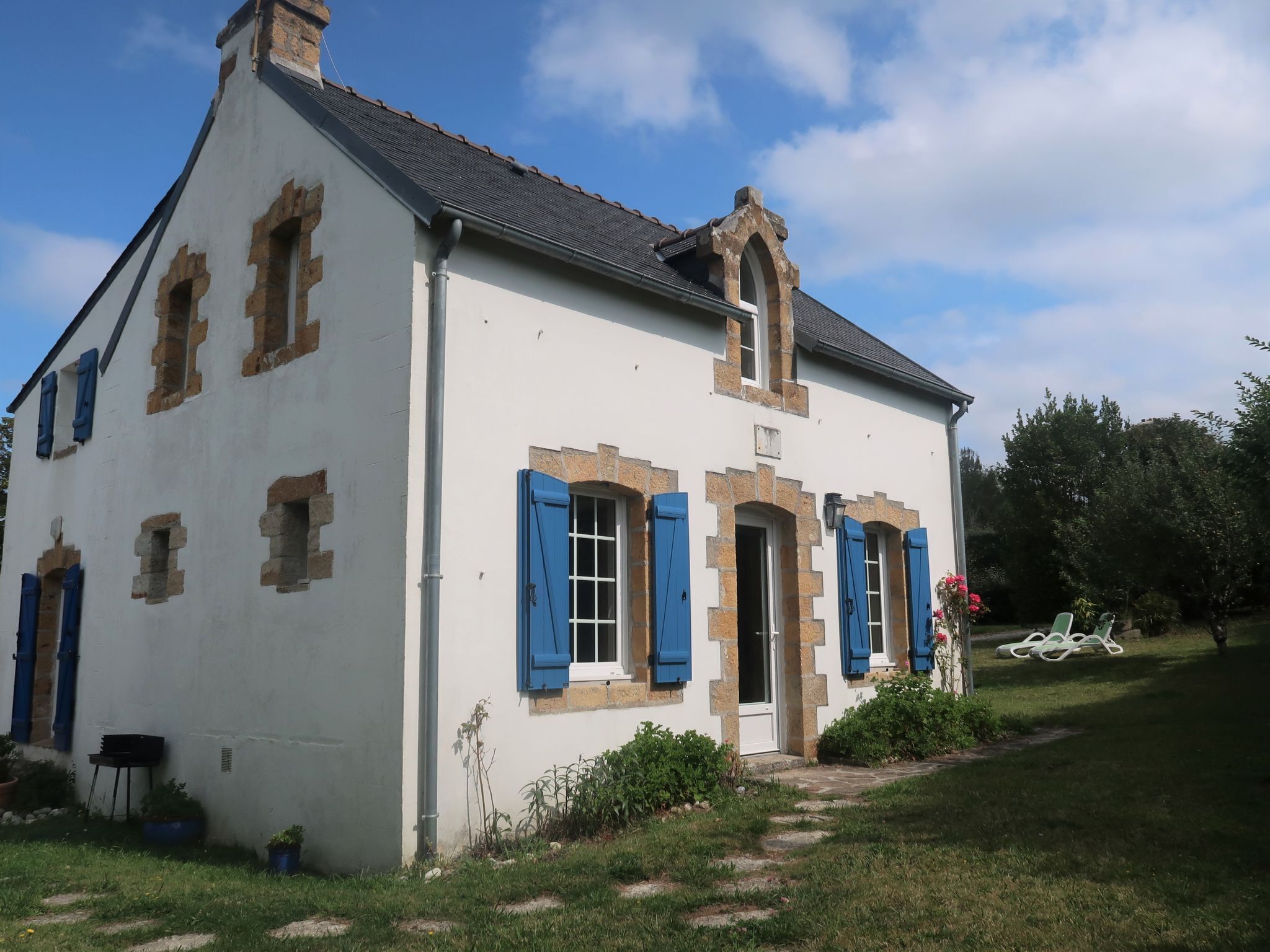 Ein weißes Gebäude mit blauen Fensterläden, einem Steinkamin und einem Gartenweg, der zum Eingang führt.