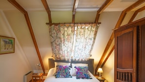Quarto com teto de madeira, cama com roupa de cama branca, cortinas florais e mesa de cabeceira com abajur.
