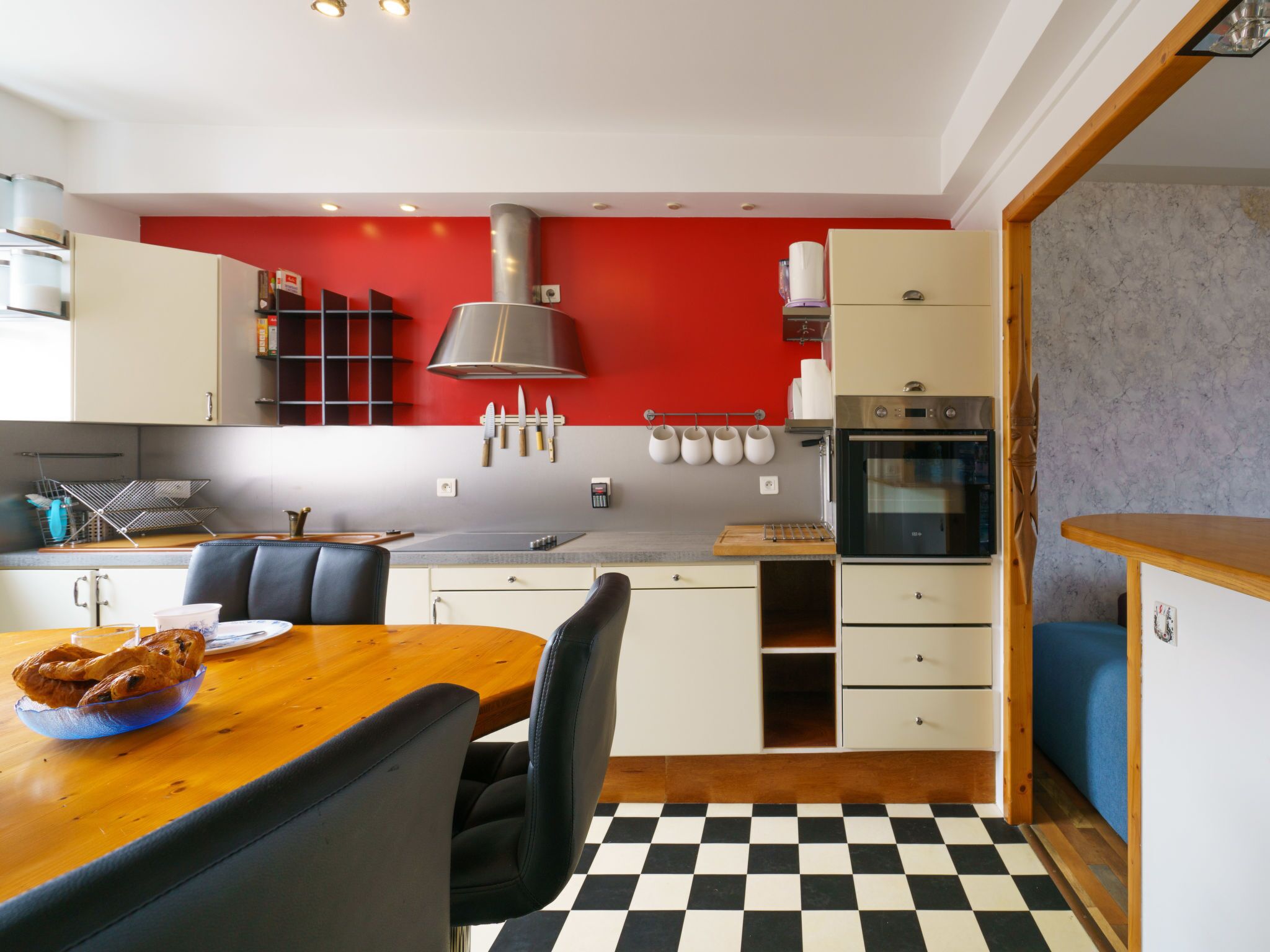 A modern kitchen with a red accent wall, a checkered floor, and a dining area with a wooden table and chairs.