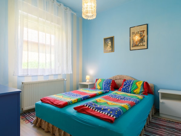 Un dormitorio con cama, ventana con cortinas, radiador, mesita de noche y decoración en la pared.