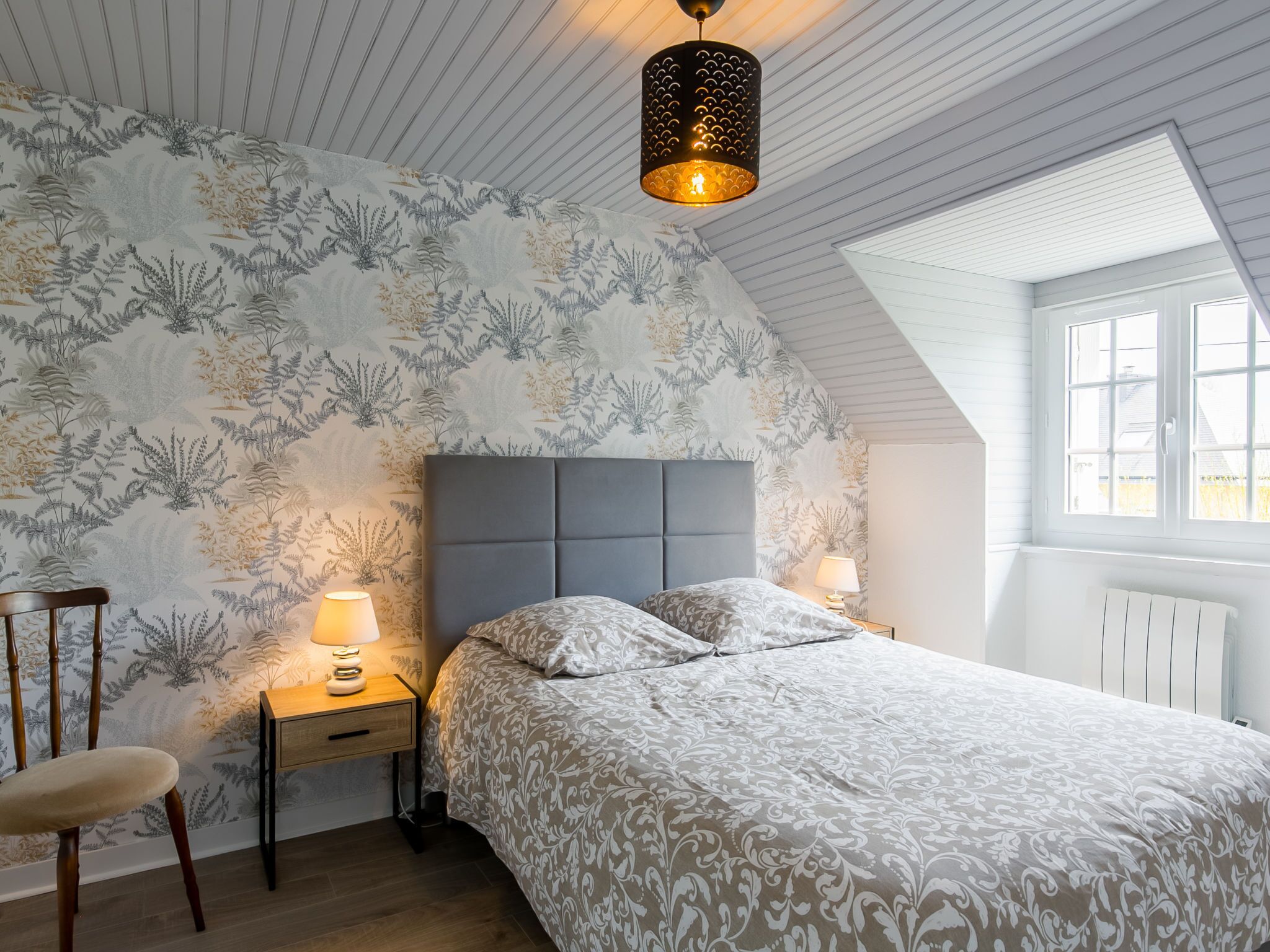 Een slaapkamer met een bed, nachtkastjes, een stoel, een hanglamp en een raam.