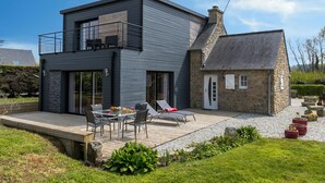A modern two-story house with a balcony, a patio with outdoor furniture, and a stone extension.