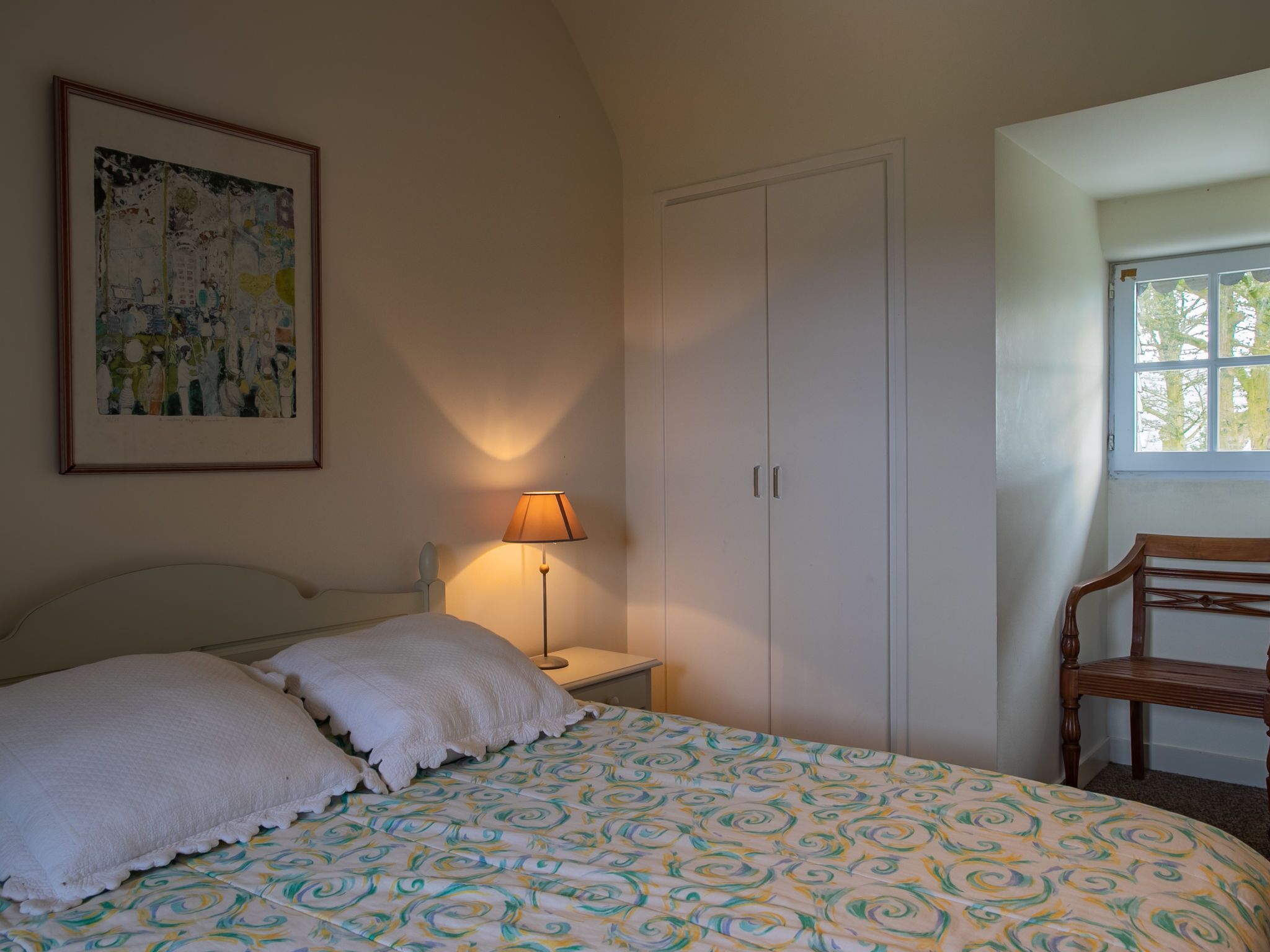 Een slaapkamer met een bed, nachtkastje, lamp, kledingkast en een raam.