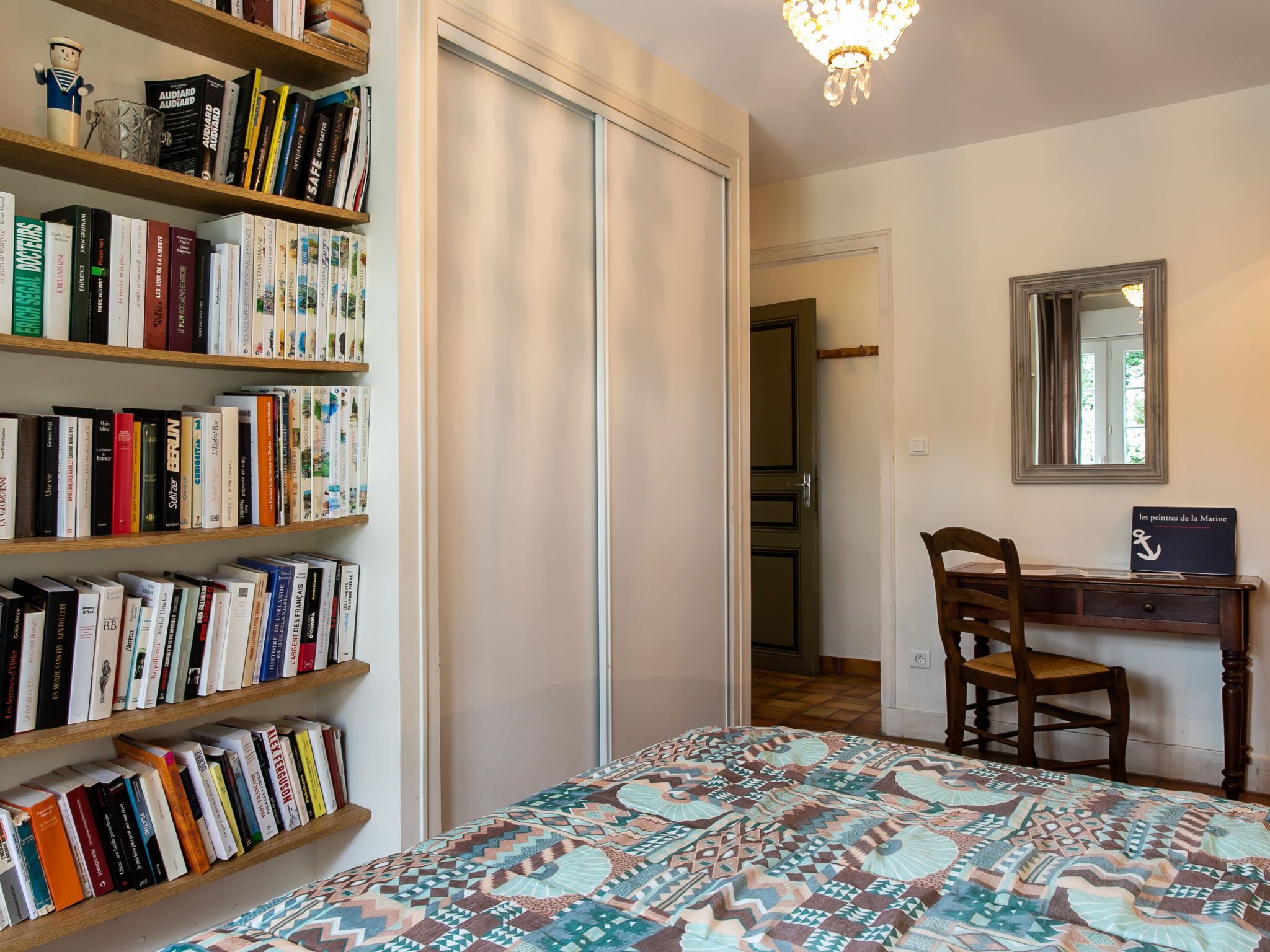 A room with a large bookshelf filled with books, a desk with a chair, a patterned bedspread, and a mirror on the wall.