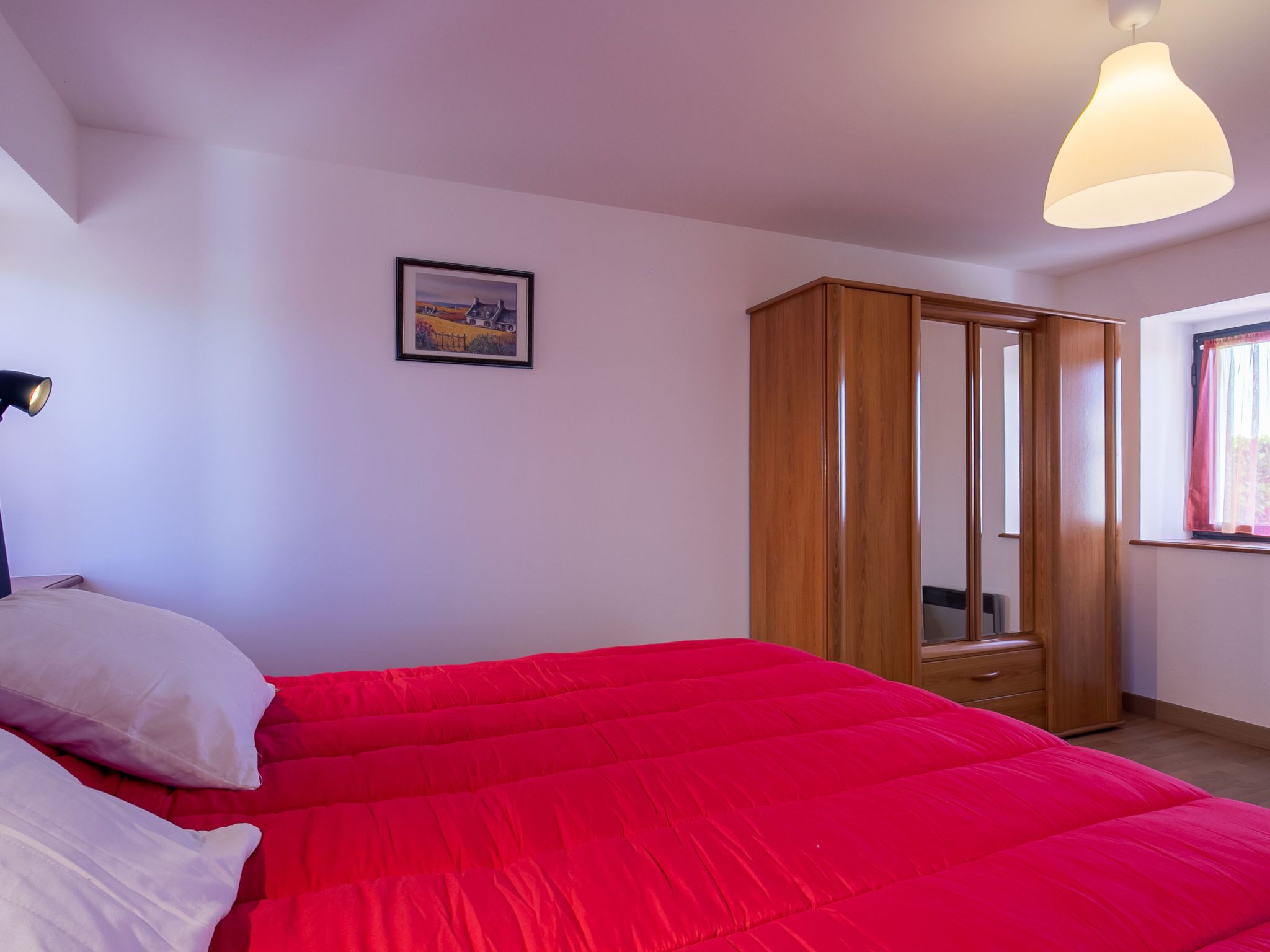 Une chambre avec un lit rouge, une armoire en bois, un tableau encadré au mur et une fenêtre avec des rideaux.
