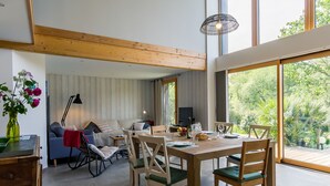 Amplia sala de estar con mesa de comedor, sillas, sofá y televisor. Amplias ventanas ofrecen vistas al verdor exterior.