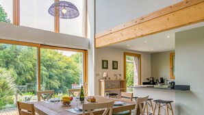 Un comedor con mesa y sillas de madera, una zona de cocina con encimera y electrodomésticos, y ventanas grandes que ofrecen vistas a zonas verdes.