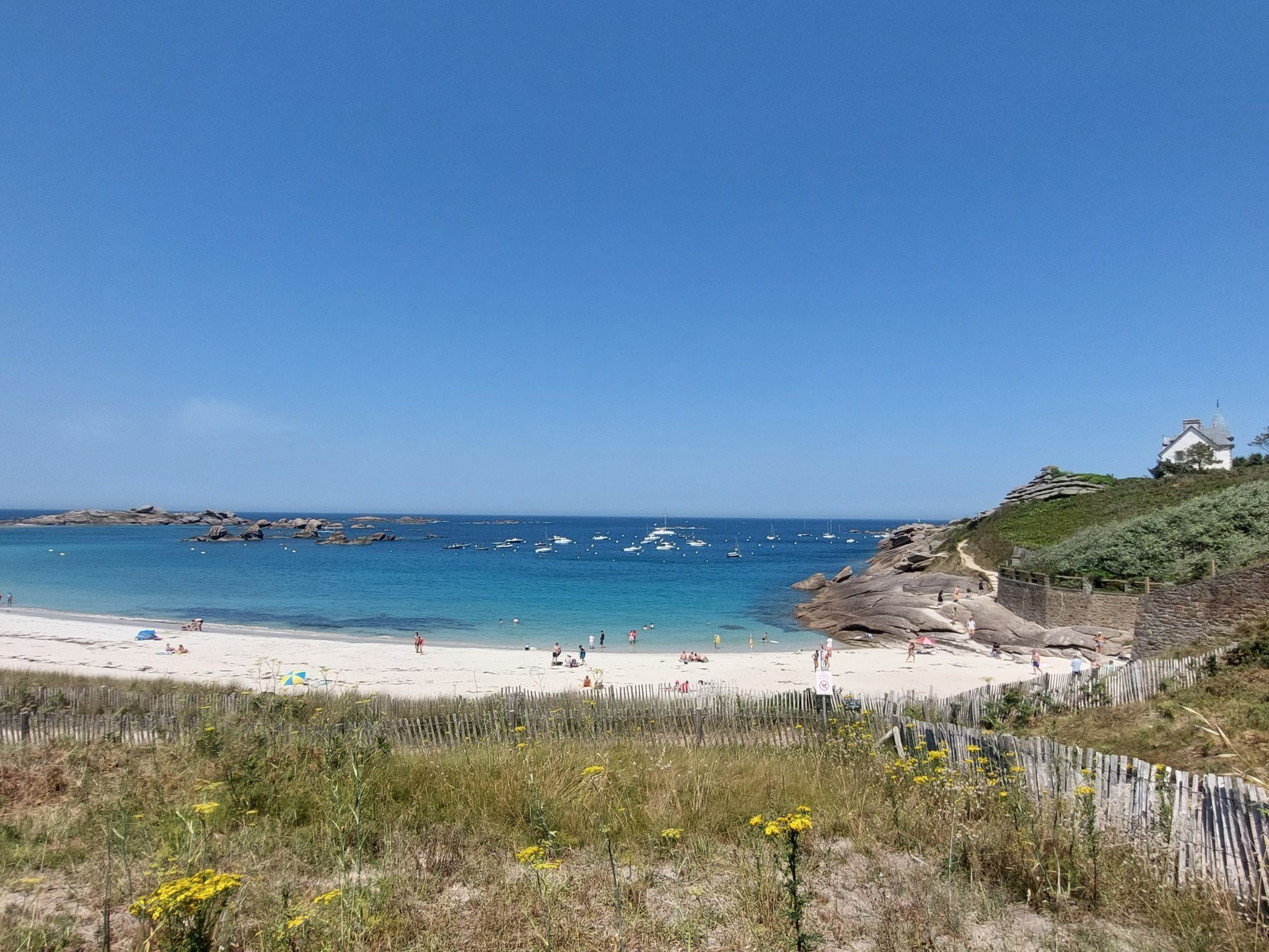 Une plage avec un ciel dégagé, des affleurements rocheux et un bâtiment blanc perché sur une colline.