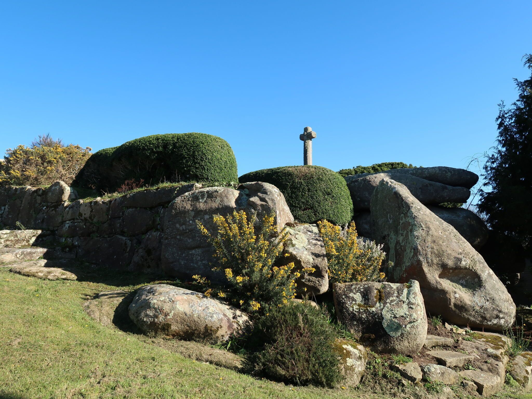 A stone cross on a grassy hill with large boulders and trimmed bushes.