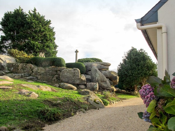 A pathway leading to a stone wall with a cross, surrounded by greenery and a building with a sloped roof. - Kerguntuil by Interhome (Pleumeur Bodou)