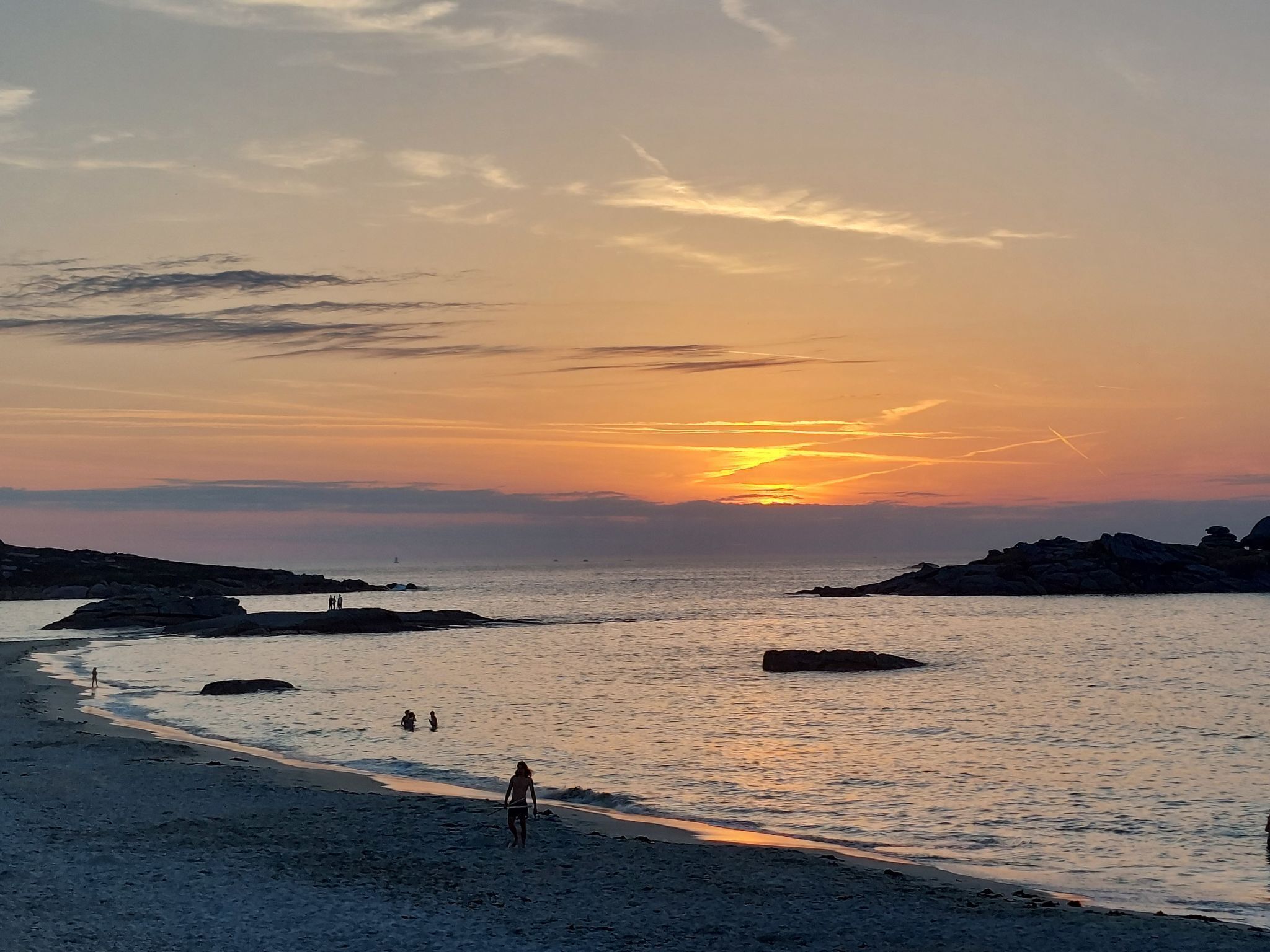 A beach scene with a sunset, rocky outcrops, and people walking along the shore.