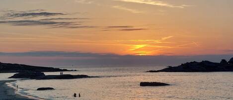 A beach scene with a sunset, rocky outcrops, and people walking along the shore.