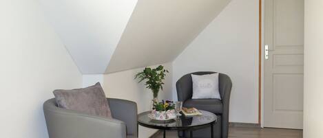A small, modern room with a sloped ceiling, a grey armchair, a round table with a plant and a book, and a grey sofa with a pillow.