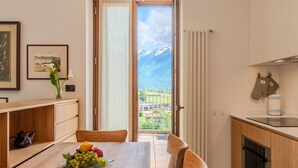 A modern kitchen with a wooden table, chairs, a fruit bowl, and a view of mountains through a sliding glass door. - Apartment Flat 1 by Interhome (Sorico Albonico)