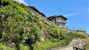 A stone staircase leading to a stone building with balconies, surrounded by lush greenery and pink flowers. - Apartment Flat 1 by Interhome (Sorico Albonico)