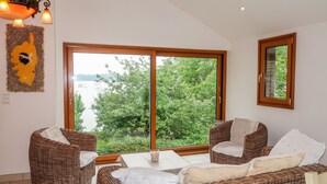 A room with wicker furniture, a large window with a view of greenery and water, and a wall-mounted light fixture. - Bella Vista by Interhome (Saint Suliac)