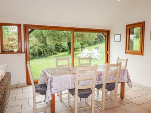 A dining area with a table and chairs, large glass doors leading to a garden, and a wicker chair to the left. - Bella Vista by Interhome (Saint Suliac)