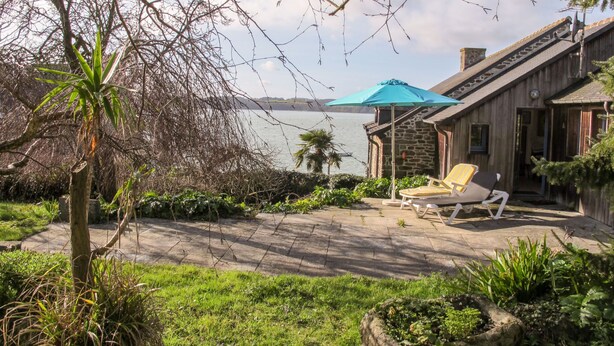 A patio with two lounge chairs and a blue umbrella, overlooking a lake with a wooden building in the background. - Bella Vista by Interhome (Saint Suliac)