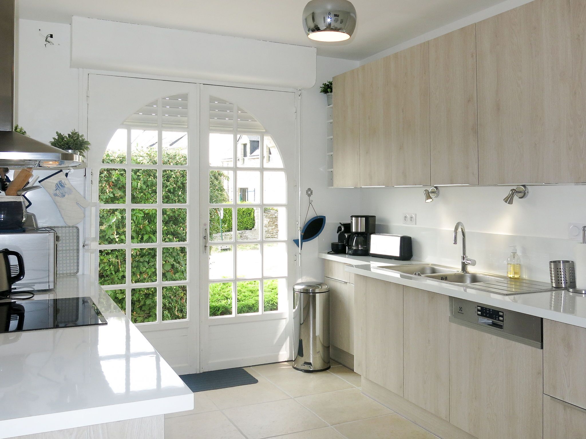 Una cocina moderna con encimera blanca, electrodomésticos de acero inoxidable y vistas a un espacio exterior verde a través de un ventanal.