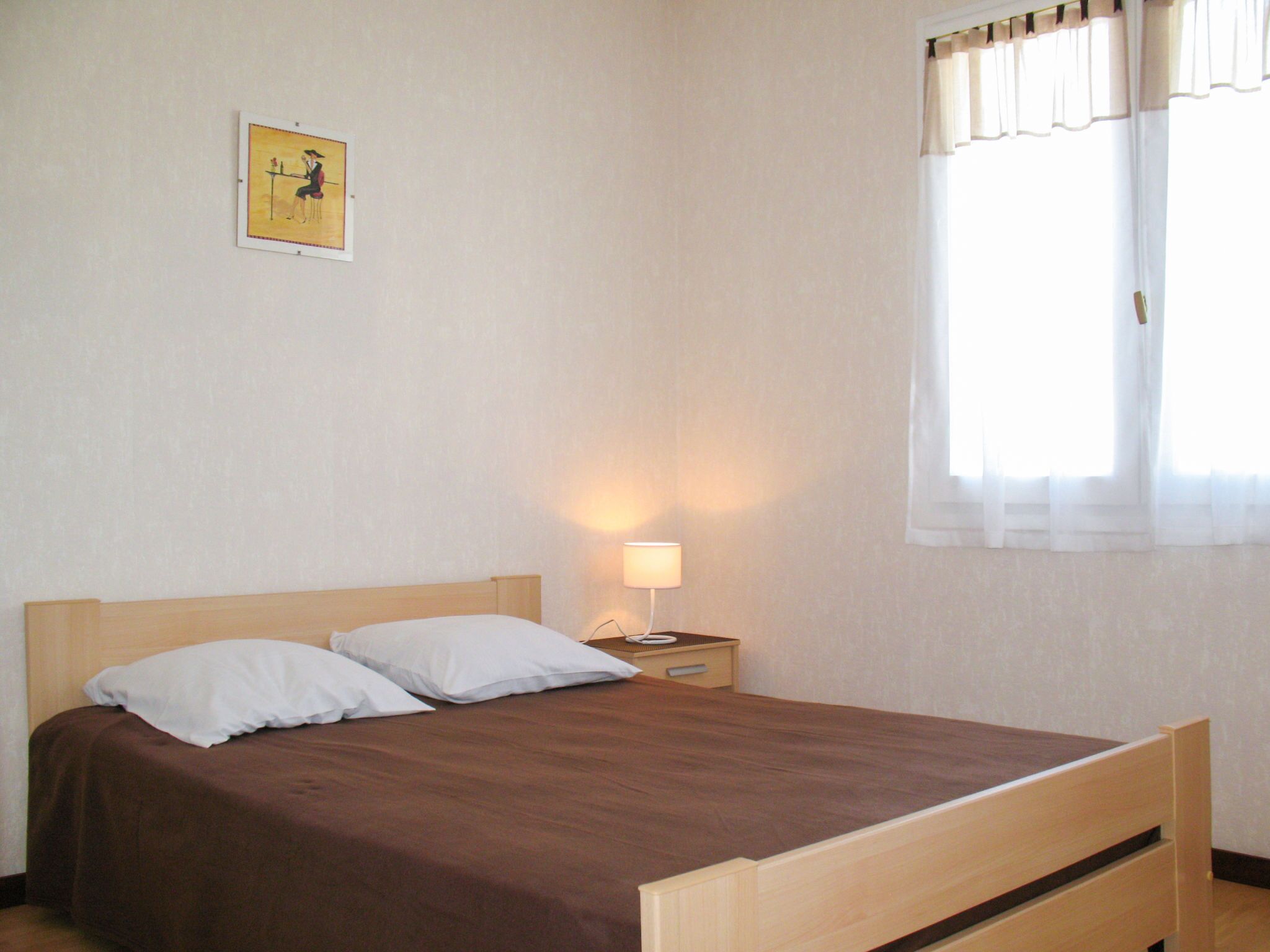 Una cama individual con estructura de madera y colcha marrón, una mesita de noche con lámpara, y una ventana con cortinas blancas.