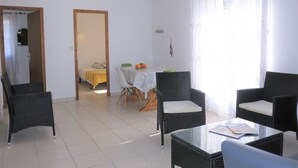 Een moderne woonkamer met rieten meubels, een eethoek met een tafel en stoelen, en een slaapkamer die door een open deur zichtbaar is.