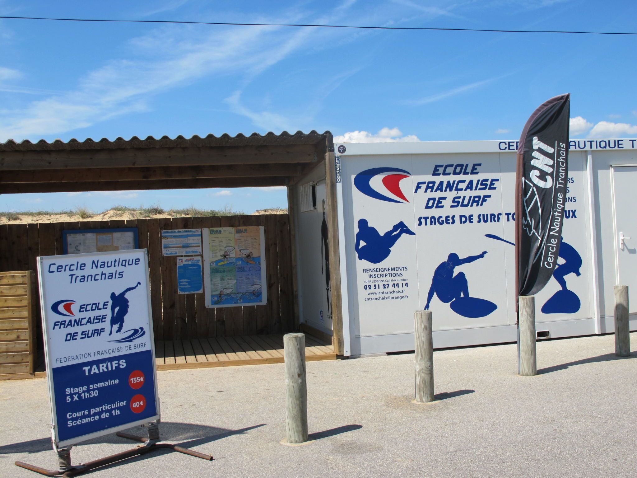 Una escuela de surf en la playa, con letreros que dicen 'Ecole Francaise De Surf' y 'Tarifas' para precios de lecciones.