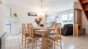 A dining area with a round table, six chairs, and a vase of flowers. There is a white refrigerator, a blue cabinet, and a window with a view of greenery. - Vacation home Néroli by Interhome (Hauteville sur Mer)