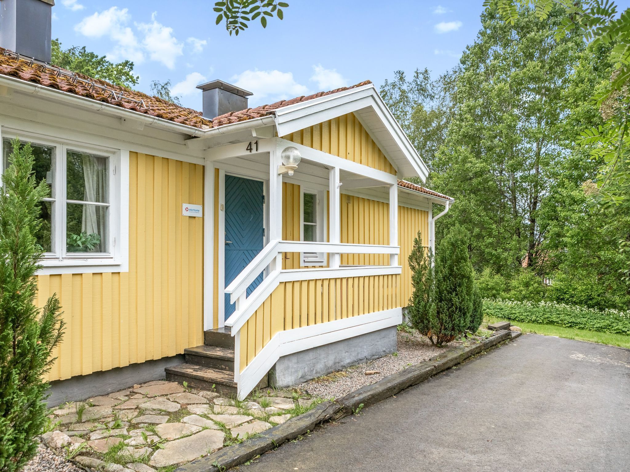 Ett gult hus med en stensatt gång som leder till entrén, en blå dörr och en liten veranda med ett vitt räcke.