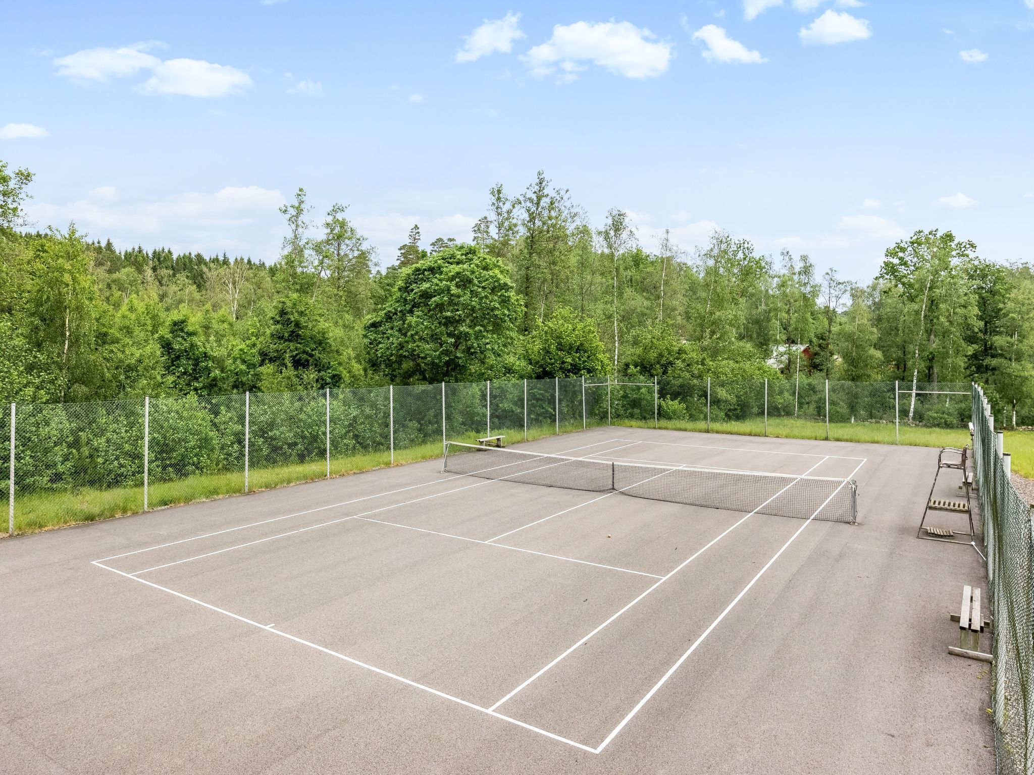An outdoor tennis court surrounded by a metal fence, with trees and a clear sky in the background.