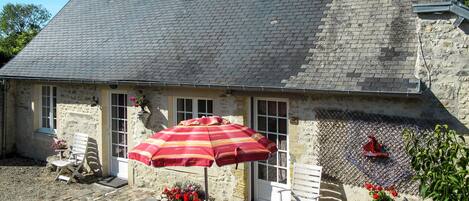 A stone building with a tiled roof, a patio with a table and chairs, and a red umbrella.