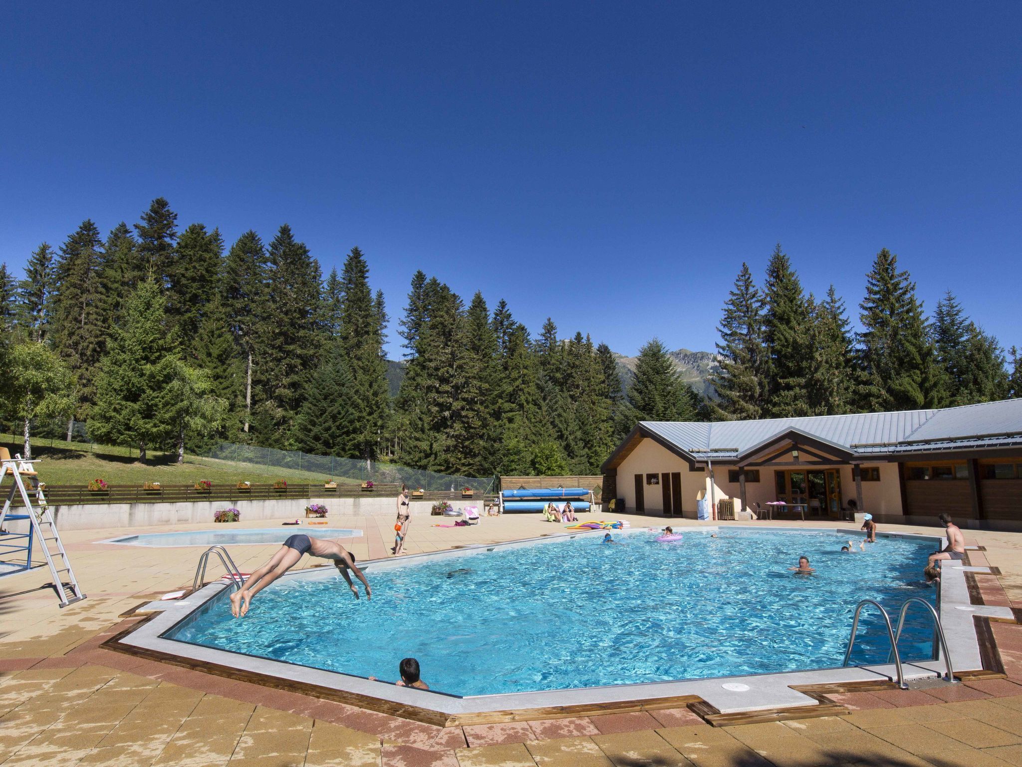 A swimming pool with people swimming and playing, surrounded by a building and trees.