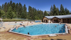 Ein Schwimmbad mit badenden und spielenden Personen, umgeben von einem Gebäude und Bäumen.