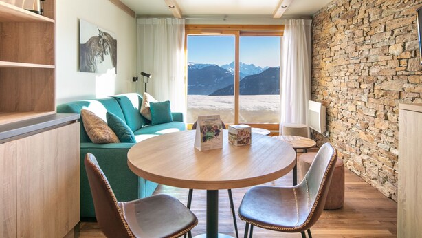 Ein gemütlicher Wohnbereich mit einem runden Holztisch, zwei Ledersesseln, einem türkisfarbenen Sofa und einer Steinwand.