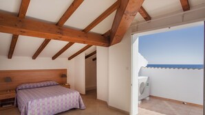 Ein Schlafzimmer mit Bett, Nachttisch, Lampe und Blick auf das Meer durch eine offene Balkontür.