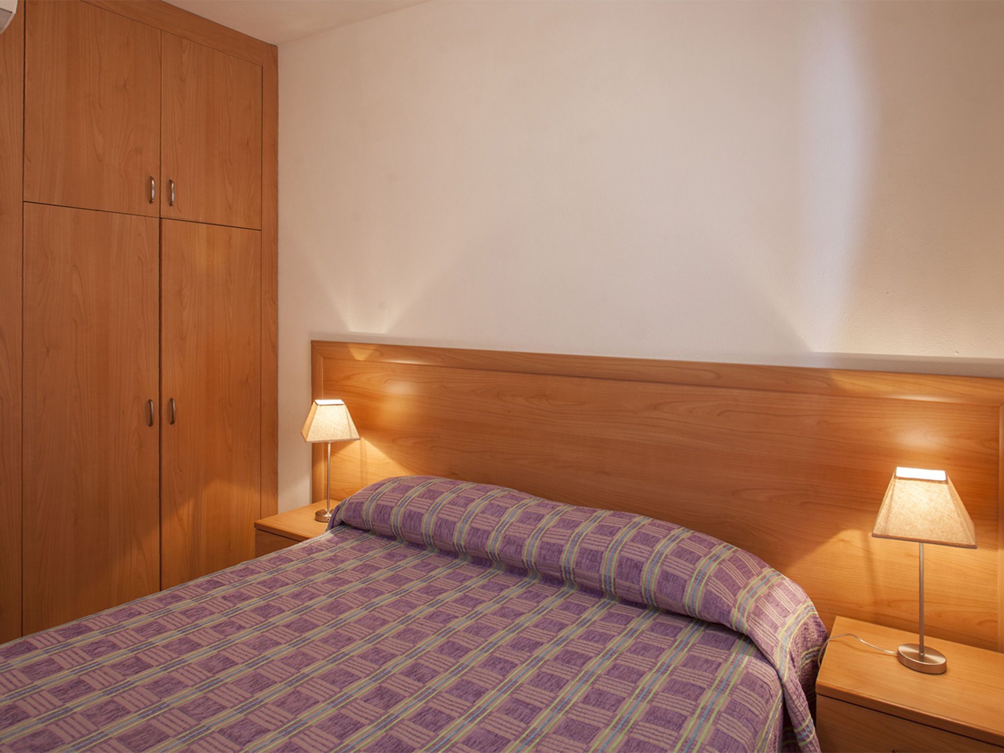 A bedroom with a wooden wardrobe, a bed with a patterned bedspread, two bedside lamps, and a wooden headboard.