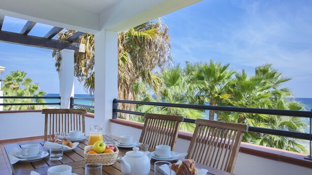 Ein Balkon mit Holztisch, gedeckt für ein Frühstück, mit Blick auf einen Strand, Palmen und das Meer.